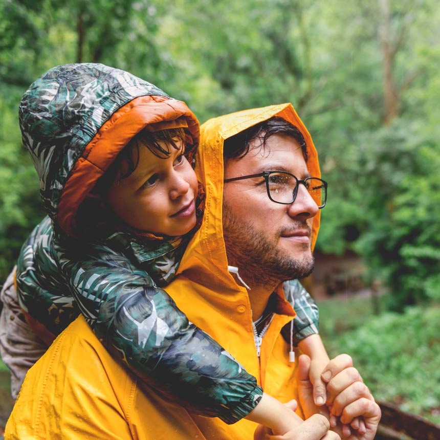 A father and son immerse themselves in the forest, wearing raincoats and calm smiles on their faces.