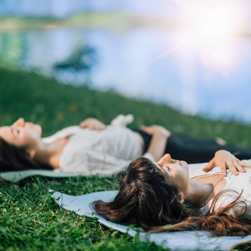 Two people lie in blankets on grass with their hands over their hearts and tummies, practicing meditation and breathing near a lake.