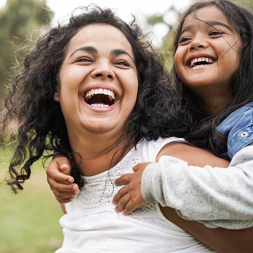 A mom walks happily through a park with her daughter on her back, both smiling.