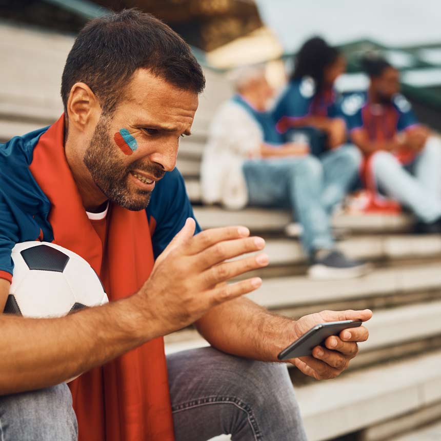 A man sits on the bleachers or steps with a soccer ball on his lap, looking at his phone with a distressed look on his face.