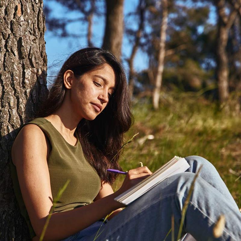 A woman sits calmly with her back on a tree trunk, writing in a journal.