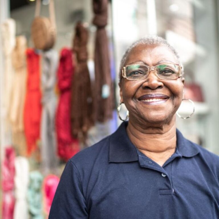 A woman with gray hair and glasses smiles at the camera, standing in front of a place with blurry colorful items in the background.