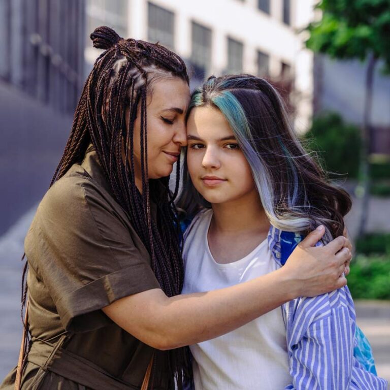 A mother holds her teenager close, while the teen looks at the camera. The teenager has a section of hair that is dyed purple and white.